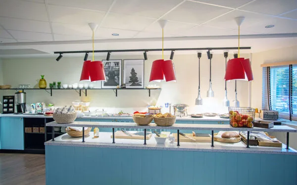 Frukostbuffé med bröd, frukt och flingor under röda lampor.