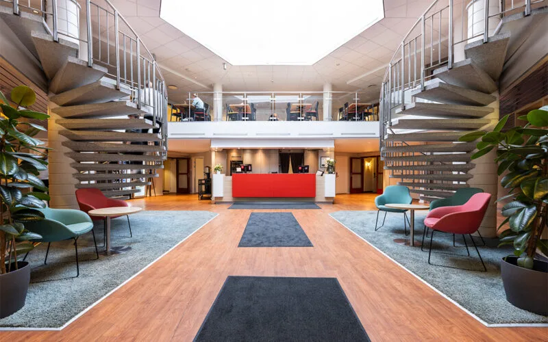 Hotellobby med trägolv, röd receptionsdisk och sittgrupper på båda sidor.