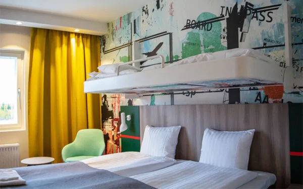 Hotellrum med dubbelsäng, extrasäng ovanför och gul gardin vid fönstret.