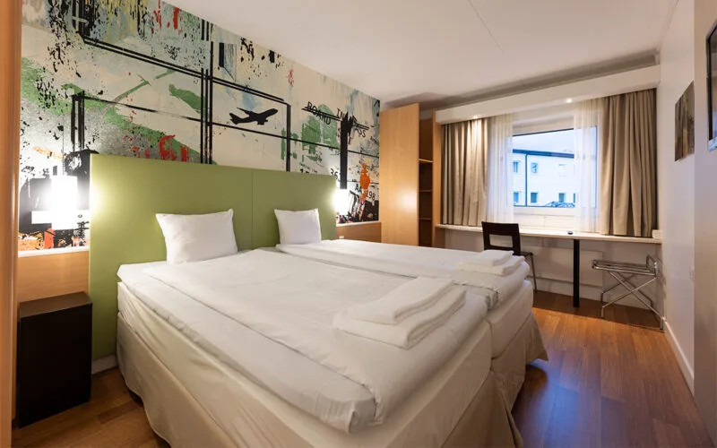 Hotellrum med bäddad dubbelsäng, skrivbord och fönster.