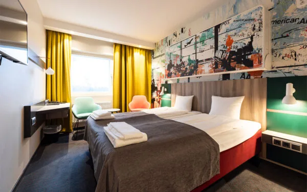 Hotellrum med dubbelsäng, färgglada fåtöljer och stora fönster med gula gardiner.