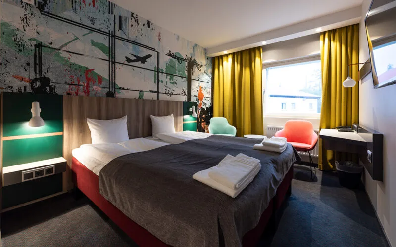 Hotellrum med två bäddade sängar, färgglada fåtöljer och gula gardiner.