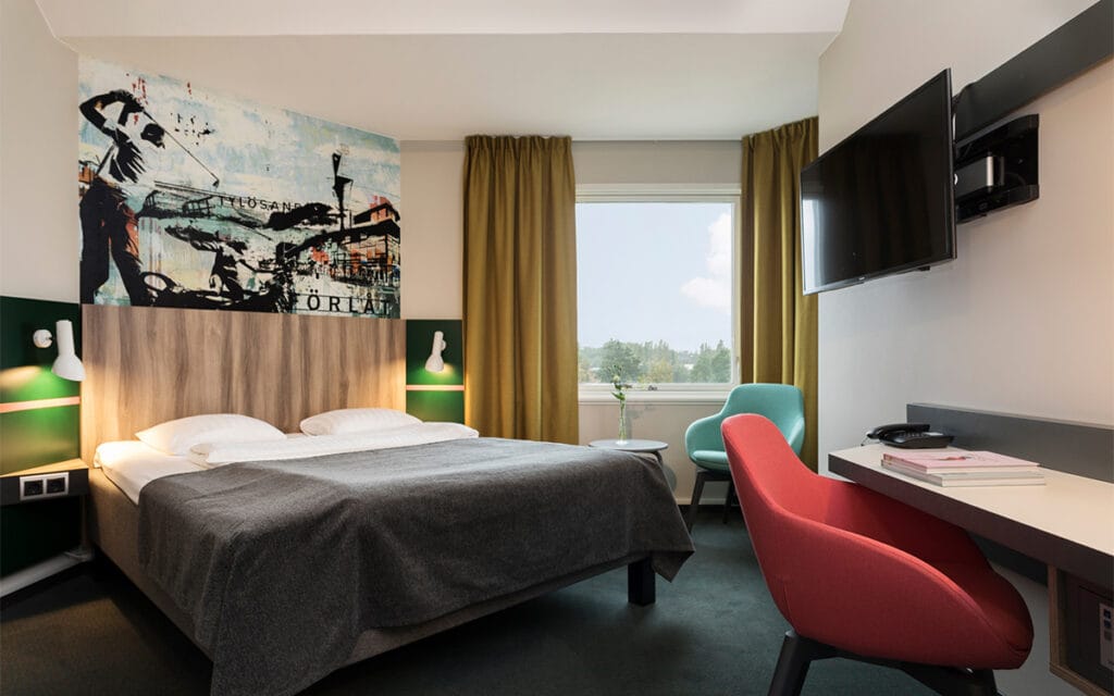 Hotelværelse med dobbeltseng, farverigt vægmaleri og gult gardin.