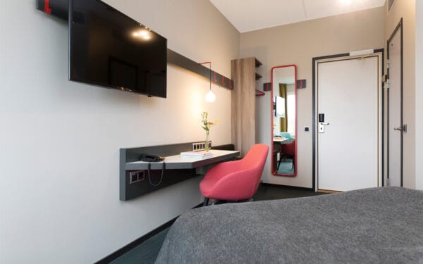 Hotellrum med dubbelsäng, skrivbord och röd fåtölj.