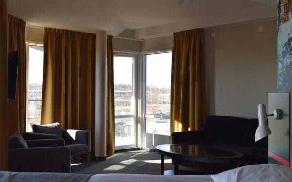 Hotellrum med sittgrupp, soffa och stora fönster med gula gardiner.
