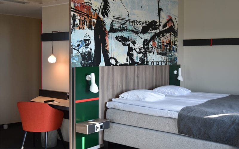 Hotellrum med dubbelsäng, färgglad väggmålning och skrivbord med röd stol.