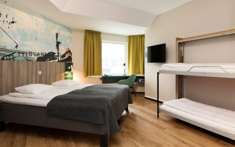 Hotellrum med dubbelsäng, våningssäng och stora fönster med gula gardiner.