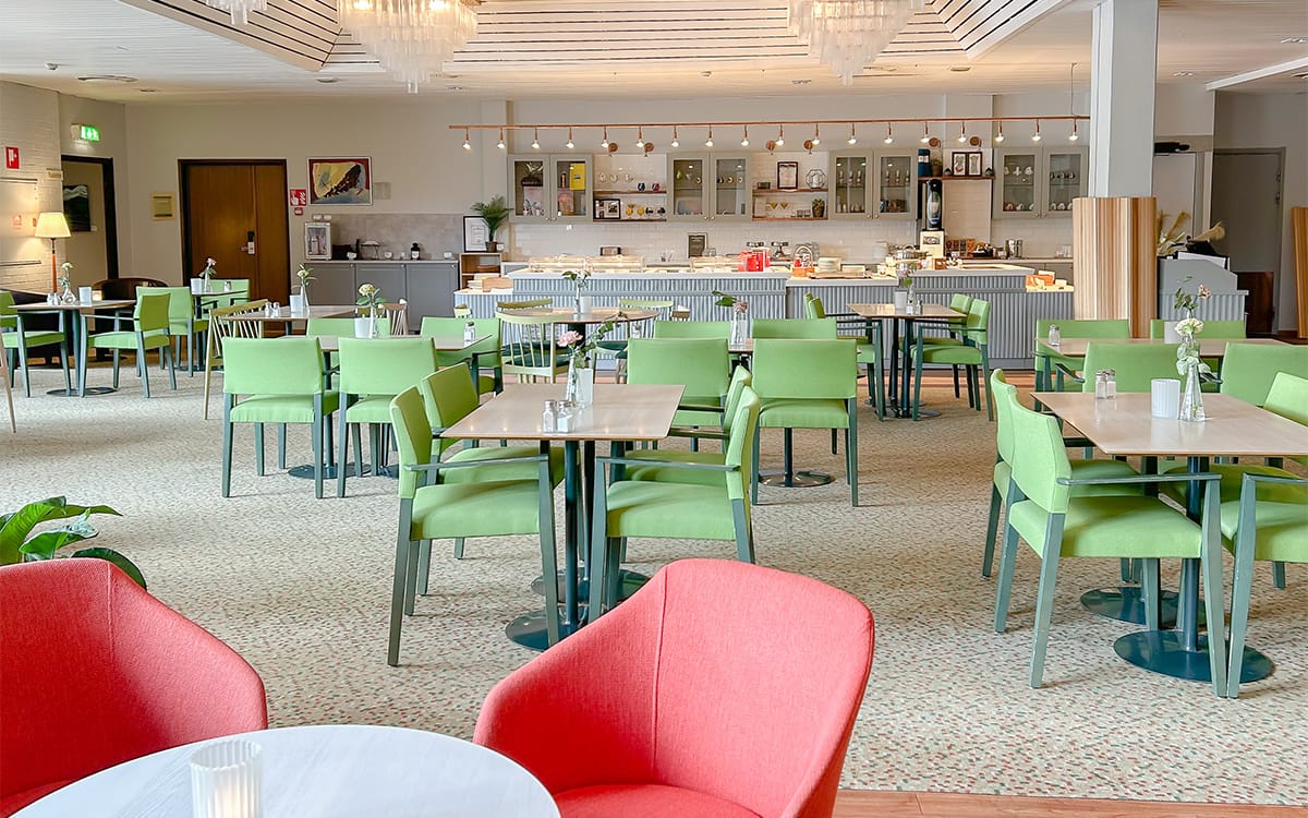 Modern and bright restaurant with green chairs and breakfast buffet in the background.