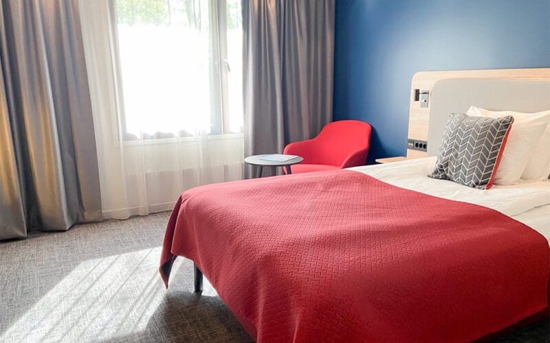 Hotellrum med enkelsäng, röd filt, blå vägg och röd fåtölj vid fönster.