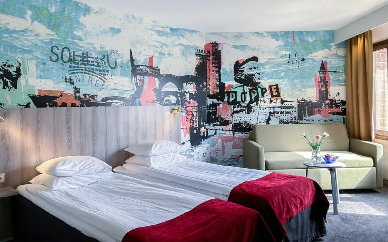 Hotellrum med två enkelsängar, soffa och färgglad väggmålning.