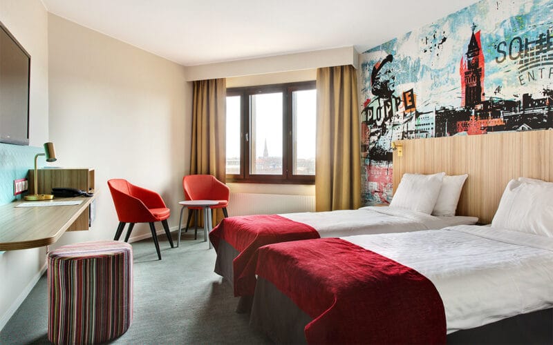 Hotellrum med två enkelsängar, röda fåtöljer och väggmålning ovanför sänggaveln.