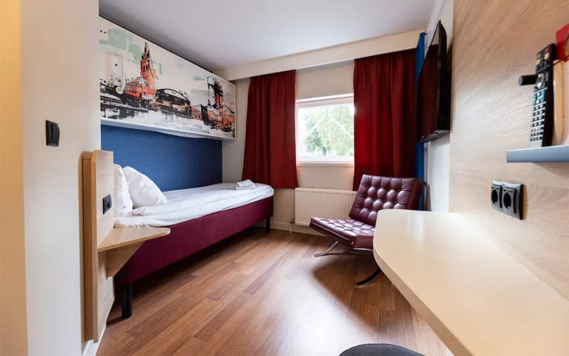 Hotellrum med två sängar, röda gardiner och fåtölj vid fönster.
