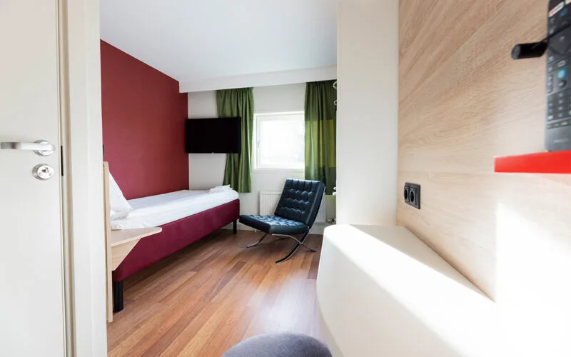 Hotellrum med säng, skrivbord och fåtölj vid fönster.