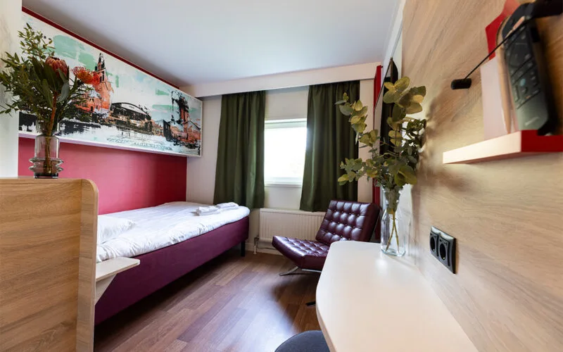 Hotellrum med enkelsäng, grön gardin, skrivbord och väggdekoration ovanför sängen.