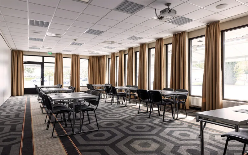 Modernt konferensrum med flera mindre bord och stolar placerade i grupper. Rummet har stora fönster längs ena väggen med långa gardiner som släpper in dagsljus.