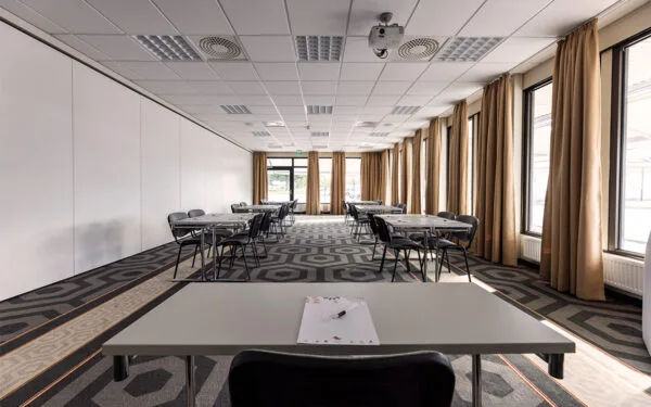 Konferensrum med småbord placerade i grupper, stora fönster med bruna gardiner längs ena väggen.