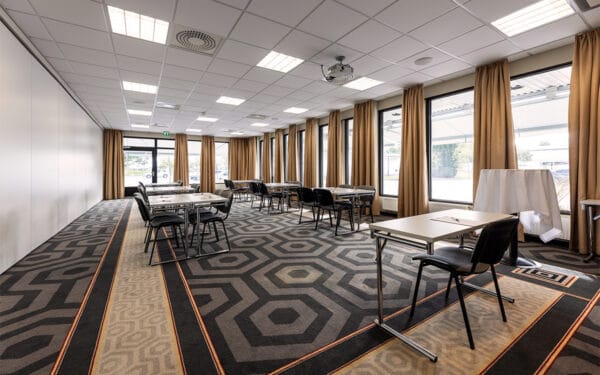 Ljust konferensrum med fönster längs hela långsidan, utrustat med bord och stolar placerade i mindre grupper.