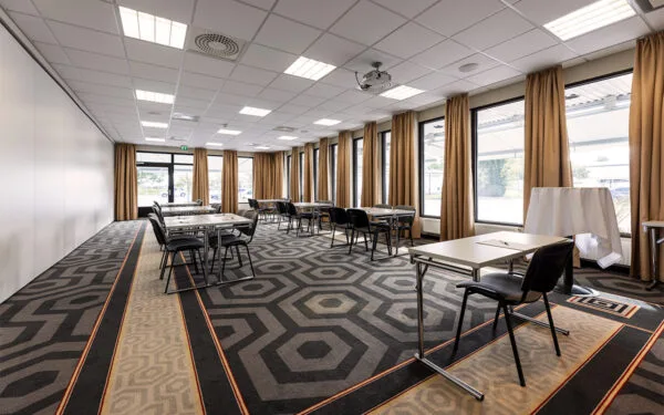 Ljust konferensrum med fönster längs hela långsidan, utrustat med bord och stolar placerade i mindre grupper.