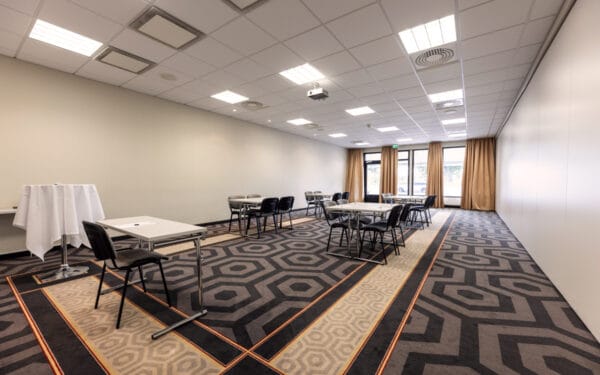 Konferensrum med småbordsmöblering och stolar utplacerade i grupper. Rummet har stora fönster med bruna gardiner, ljusa väggar, projektor i taket och en heltäckningsmatta med geometriskt mönster.