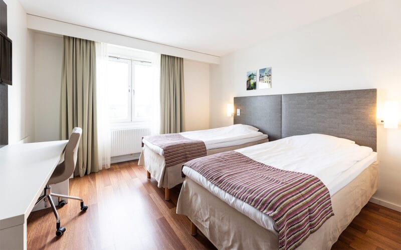 Hotellrum med två enkelsängar, skrivbord med stol, TV på väggen och stort fönster med gardiner.