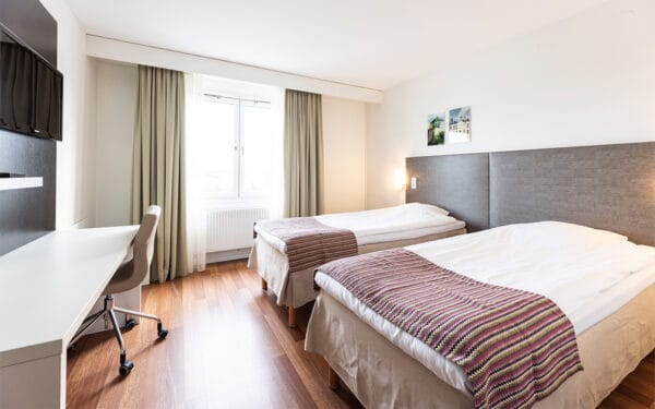 Hotellrum med två enkelsängar, skrivbord med stol, TV på väggen och stort fönster med gardiner.