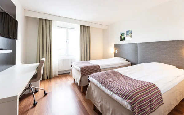 Hotellrum med två enkelsängar, skrivbord med stol, TV på väggen och stort fönster med gardiner.