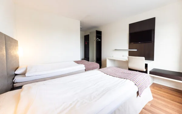 Hotellrum med två enkelsängar, skrivbord med stol, TV på väggen och öppen garderob.