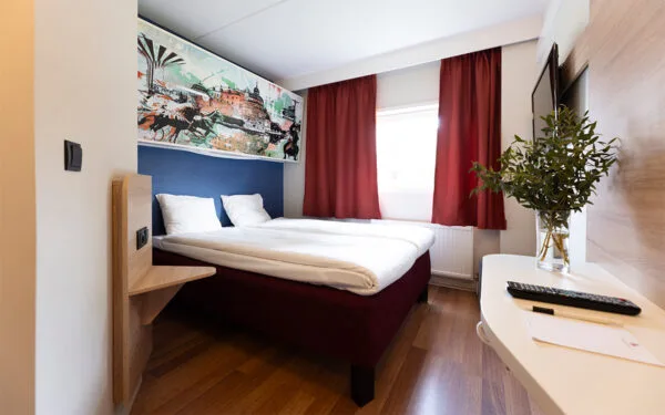 Hotellrum med dubbelsäng, överslaf, röda gardiner och TV på väggen.