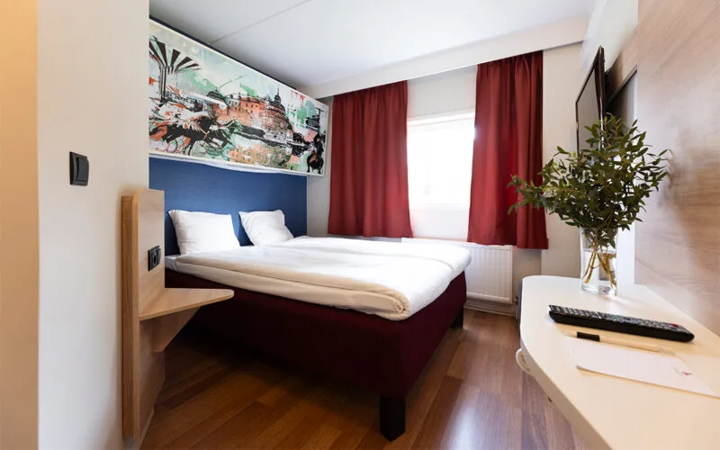 Hotellrum med dubbelsäng, överslaf, röda gardiner och TV på väggen.