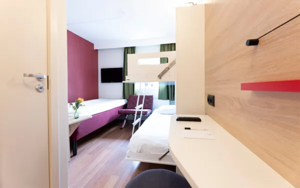Hotellrum med enkelsäng, våningssäng, skrivbord och gröna gardiner.