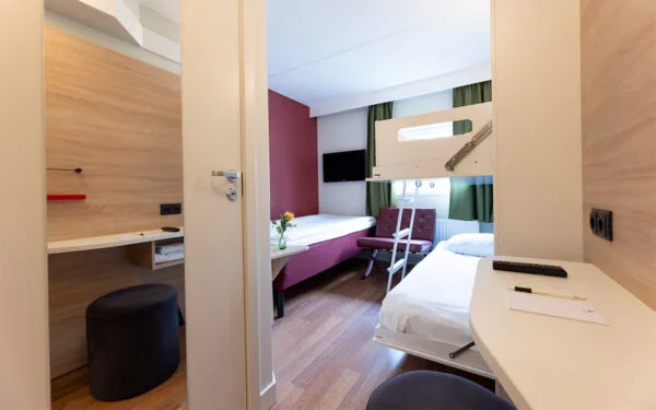 Hotellrum med enkelsäng, våningssäng, skrivbord och gröna gardiner.