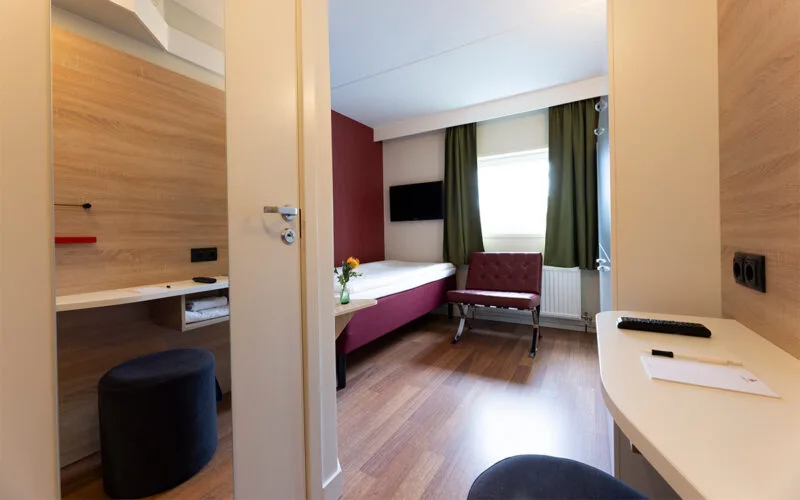 Hotellrum med enkelsäng, våningssäng, skrivbord och gröna gardiner.