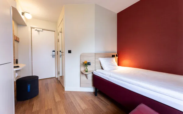 Hotellrum med enkelsäng, röd fondvägg och stängd utfällbar våningssäng i hörnet av bilden.