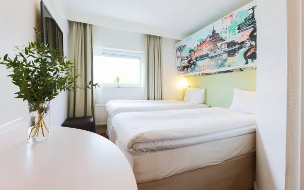 Hotellrum med dubbelsäng, ljusa gardiner, TV på väggen och en väggmålning ovanför sängen.