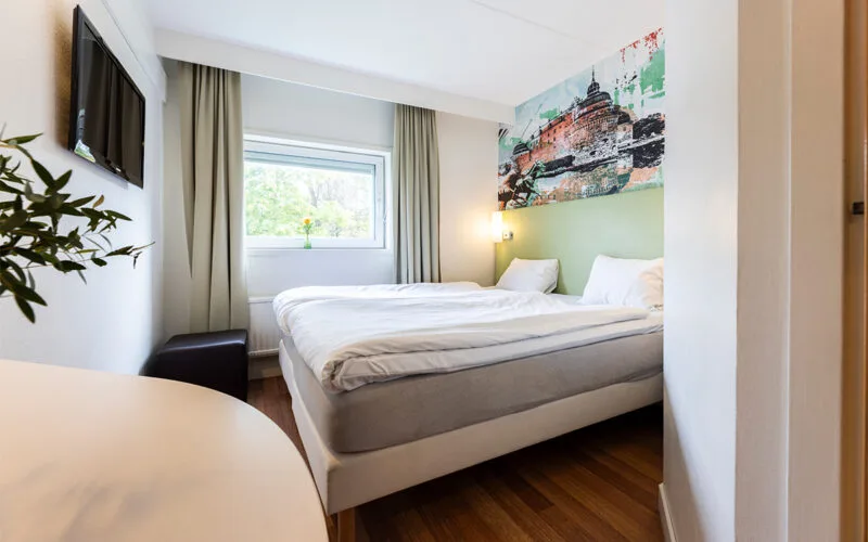Hotellrum med dubbelsäng, ljusa gardiner, TV på väggen och en väggmålning ovanför sängen.