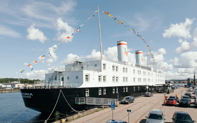 Hotellfartyget målat i svart och vitt med två skorstenar, ligger förtöjt vid kajen. Färgglada flaggor är hissade längs en lina över däck och bilar står parkerade längs gatan intill.