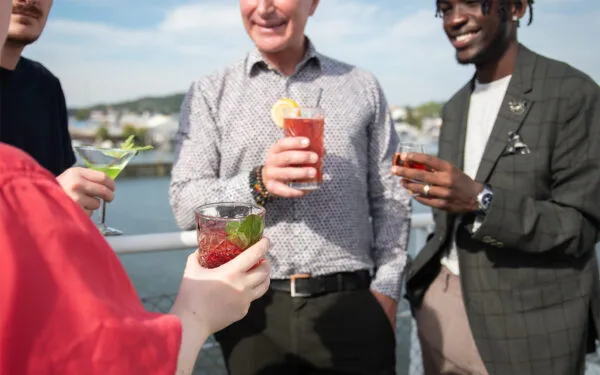 Fyra personer står på däck och håller färgglada drinkar i händerna under en social stund vid vattnet.