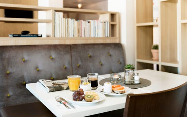 Frukostbord dukat med tallrikar innehållande bröd, frukt, yoghurt och croissant samt glas med juice och kaffe, placerat i en matsal med bokhyllor och stoppad bänk i bakgrunden.