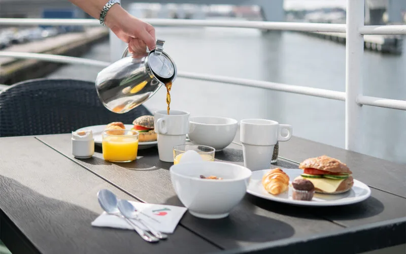 Frukost serverad på ett bord utomhus på däck, med kaffe som hälls upp ur kanna, juice, bröd, croissant, yoghurt och muffins.