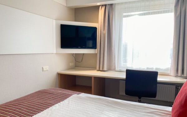Hotellrum med säng, skrivbord, skrivbordstol, TV och fönster.