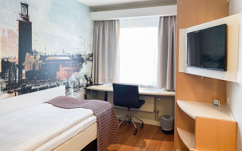 Hotellrum med enkelsäng, skrivbord vid fönster och väggmålning ovanför sängen.