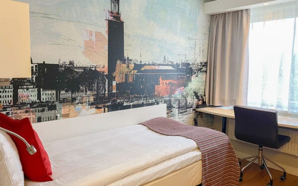 Enkelrum med säng, skrivbord vid fönster och väggmålning.