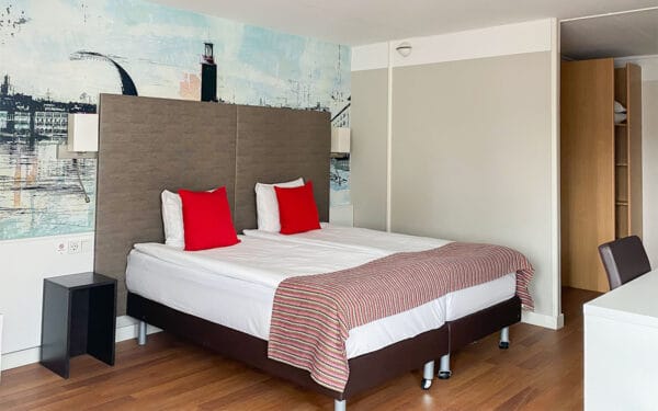 Hotellrum med dubbelsäng, grå sänggavel, röda kuddar och trägolv.