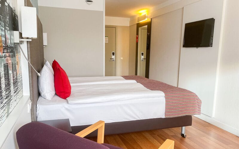 Hotellrum med två bäddar, röd kudde, tv på väggen och fåtölj vid sidan.