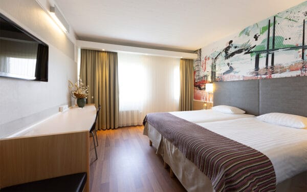 Hotellrum med dubbelsäng, skrivbord och stora fönster.