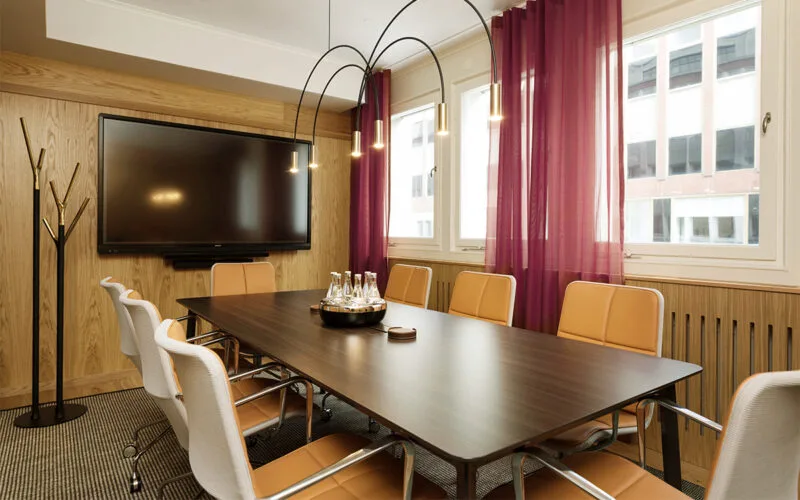 Modernt konferensrum med långbord, bekväma stolar, stora fönster med lila gardiner och en stor skärm på väggen