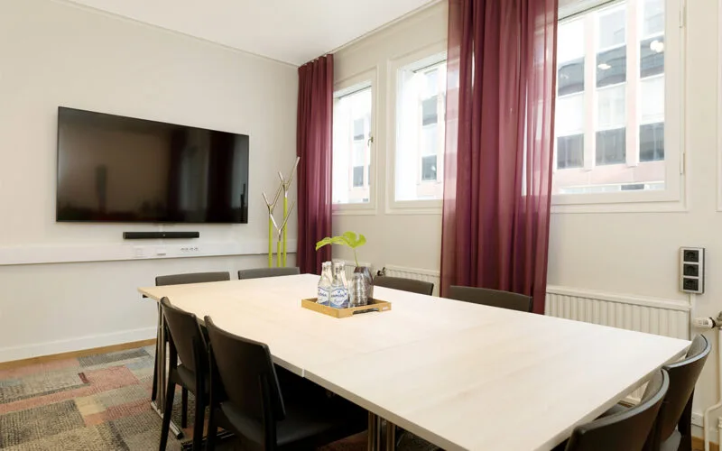 Ljust mötesrum med stort bord, svarta stolar, stora fönster med lila gardiner och en väggmonterad TV-skärm