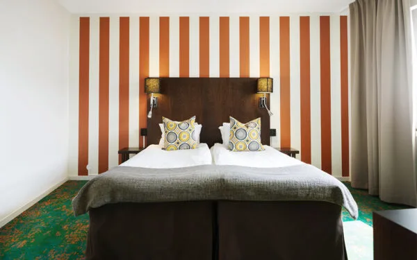 Hotellsäng med två kuddar framför fondvägg med breda orangea ränder