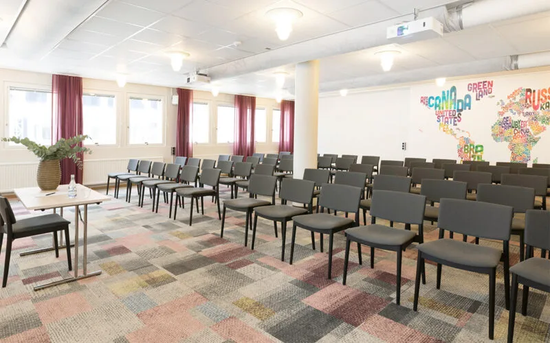 Konferensrum med rader av svarta stolar, färgglatt mönstrad heltäckningsmatta och stora fönster med röda gardiner.