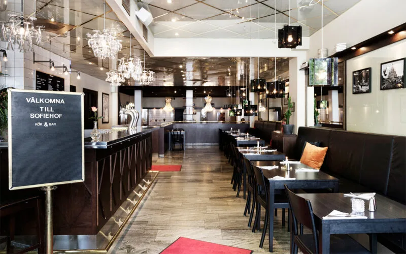 Restauranginteriör med mörka trädetaljer, lång bardisk, kristallkronor i taket och dukade bord längs väggarna